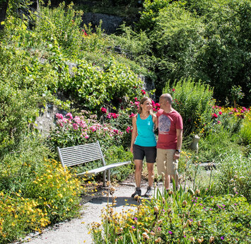 Ein Mann und eine Frau laufen lächelnd durch eine Gartenanlage mit vielen bunten Blumen.