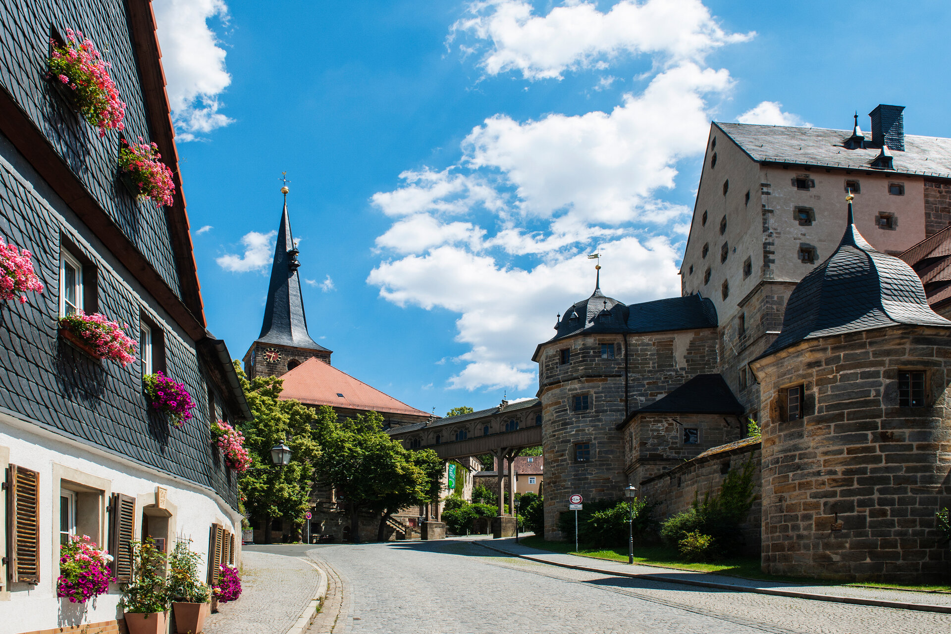 Auf der linken Seite steht ein Schieferverkleidetes Haus. Im Hintegrund eine Kirche, von der ein Gebäudebrücke aus Holz zum Schloss aus Sandstein führt.
