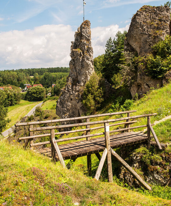 Im Vordergrund verbindet eine hölzerne Brück zwei grüne Hügel. Im Hintergrund ist eine hochaufragende Felsnadel.